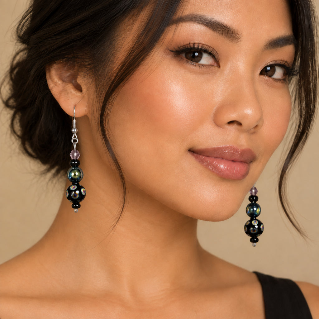 A pair of handmade dangle earrings with silver fishhook wires, small metallic beads, and glossy black glass beads featuring iridescent multicolor patterns, displayed on a woman