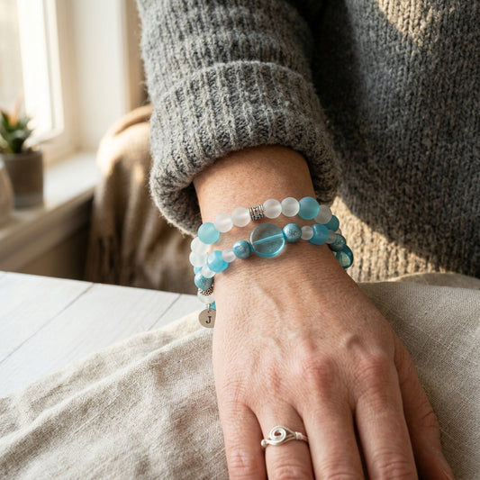 Two beaded stretch bracelets displayed on a light wood plank background. The top bracelet features translucent blue and white round beads with a small silver charm.