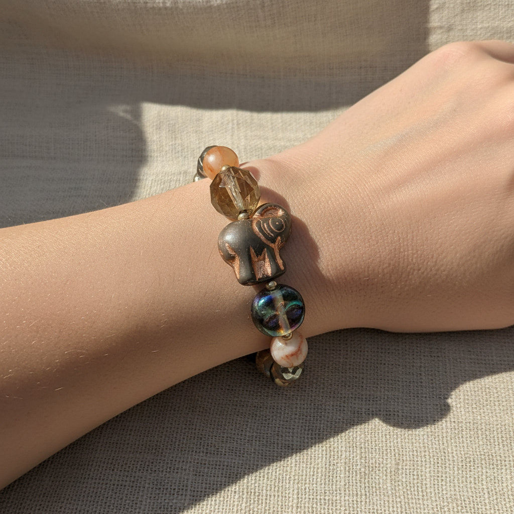 A beaded stretch bracelet featuring a carved copper elephant charm, a green stone bead, a faceted gray stone, and marbled pink and white beads, displayed on a textured beige linen background.