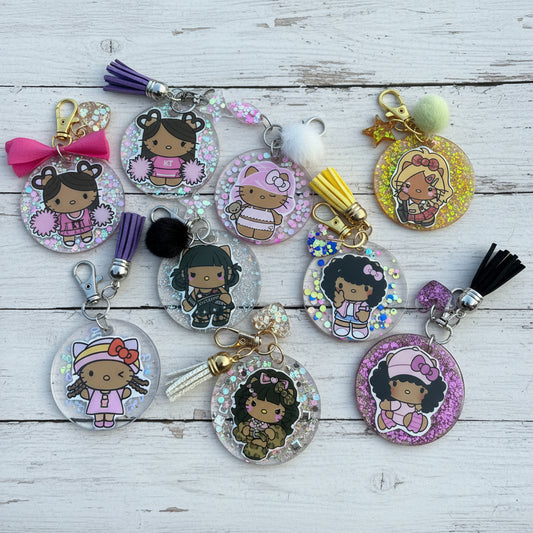 A group of round acrylic keychains featuring cartoon girl illustrations, some with glitter backgrounds and tassel charms, arranged on a white surface.