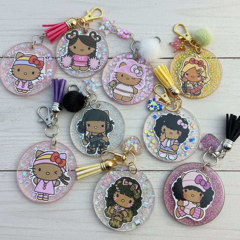 A group of round acrylic keychains featuring cartoon girl illustrations, some with glitter backgrounds and tassel charms, arranged on a tan surface.