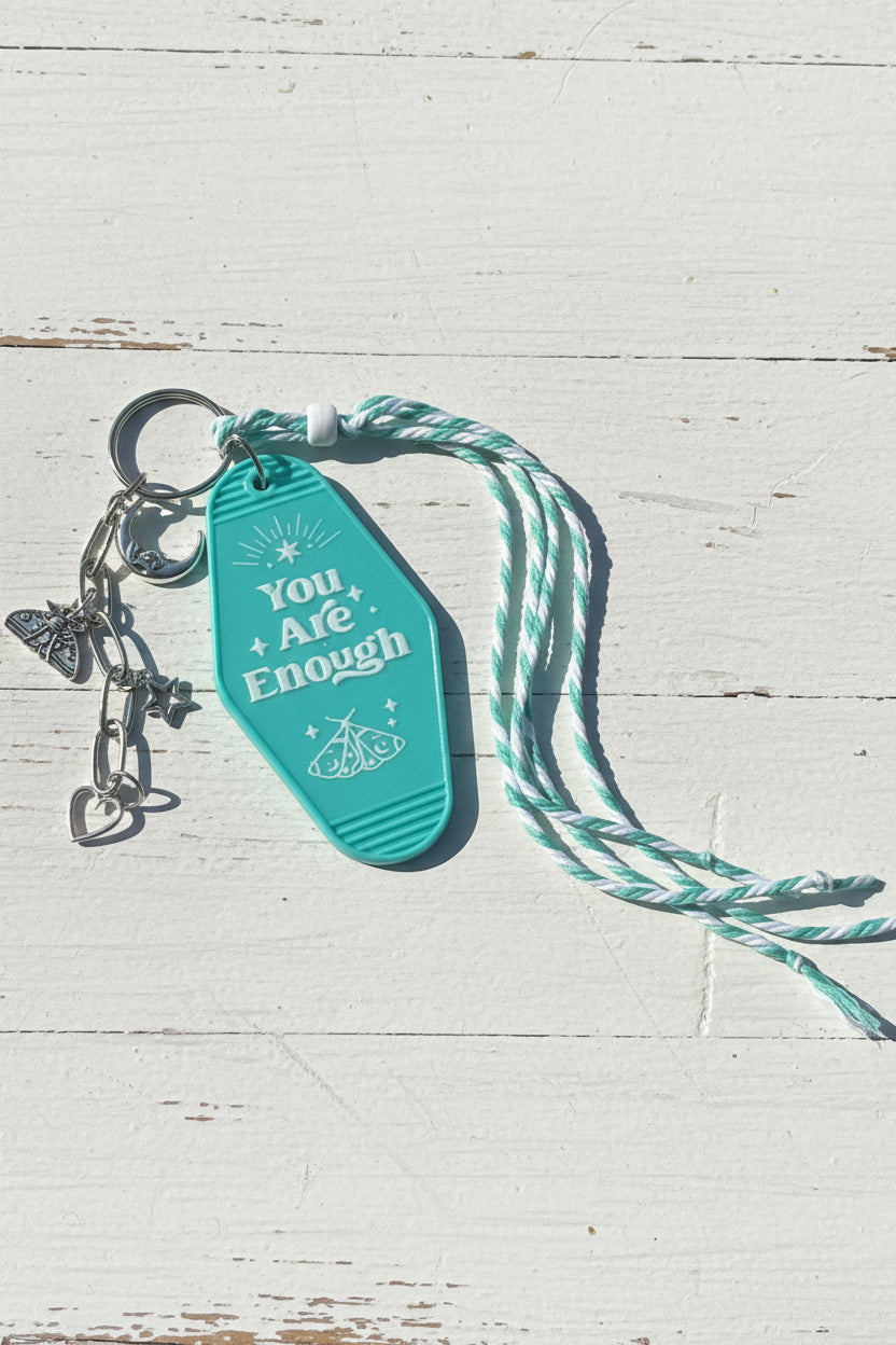 A turquoise keychain with the words 'You Are Enough' and a butterfly design, attached to a mint and white twisted rope and silver keyring, placed on a brown cardboard surface.