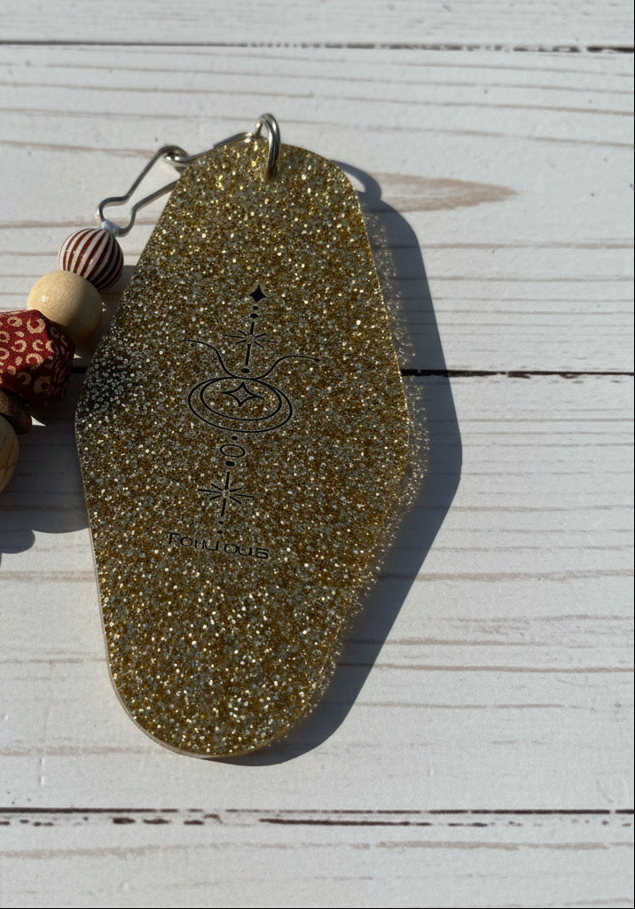 A coffin-shaped pendant covered in dense gold glitter, featuring a dark engraved symbol in the center, resting on a brown textured surface with a beaded necklace partially visible on the left.