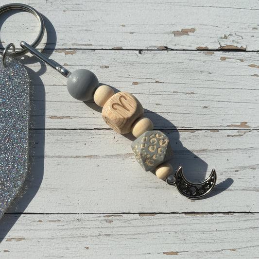 A close-up of a beaded keychain charm with wooden beads, a gray bead, a silver crescent moon charm, and a silver key ring, placed on a white background.