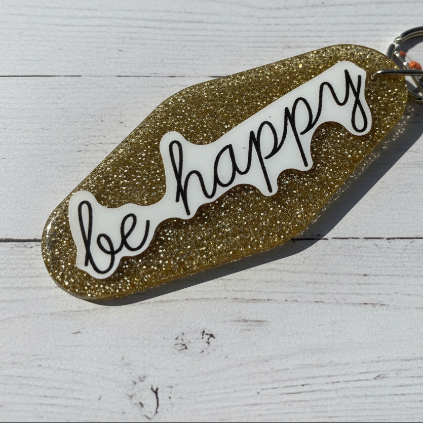 A glittery gold keychain with a white die-cut sticker that reads 'be happy' in black script, photographed on a brown textured surface.