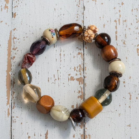 A beaded stretch bracelet featuring a mix of polished and textured stone beads in brown, orange, white, and black, with a small silver metal charm, displayed on a white background.