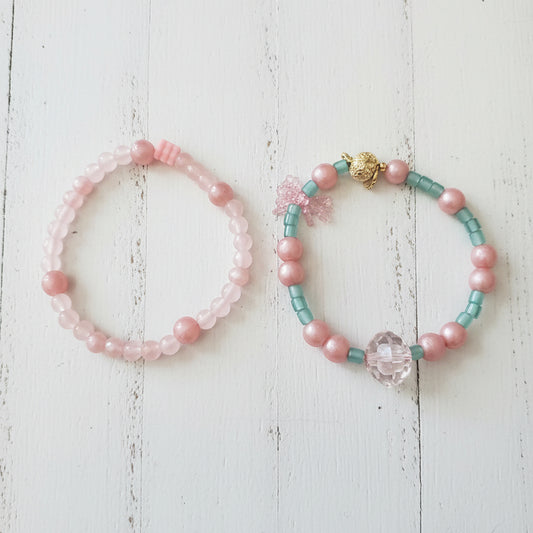 Two beaded bracelets displayed on a white wood. One bracelet is made of light pink beads, and the other is made of turquoise blue beads with a central pink heart charm.