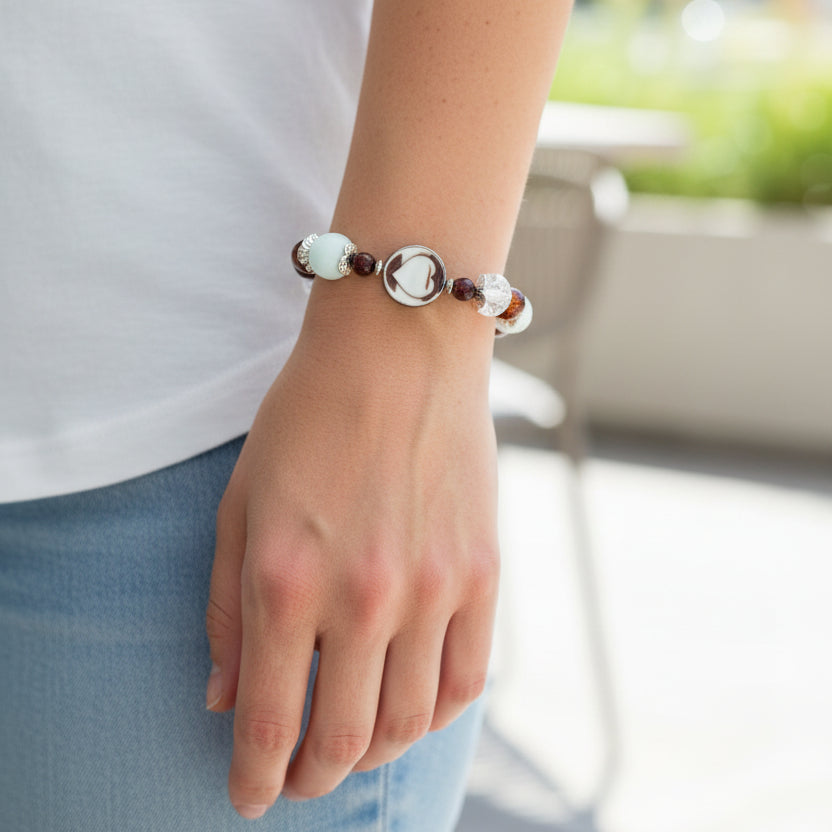 A beaded stretch bracelet featuring white, brown, and clear beads, including a prominent white heart-shaped bead, displayed on a woman's arm.