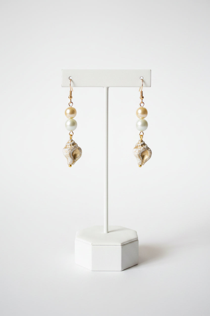 A pair of dangling earrings with gold-tone hooks, light blue beads, white pearl-like beads, and textured white and gold shell charms, displayed on a stand.