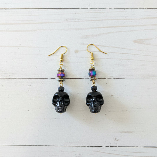 A pair of handmade dangle earrings with gold fishhook wires, small multicolored beads, and dark carved skull charms, resting on a white planks