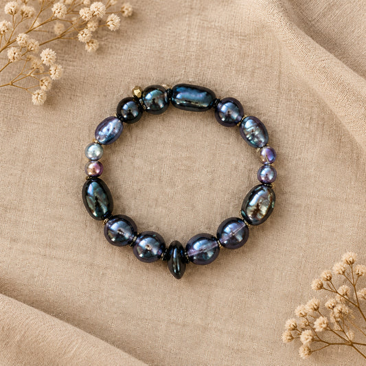 A beaded stretch bracelet featuring dark polished black and gray stones with small iridescent accents, displayed on a textured brown background.