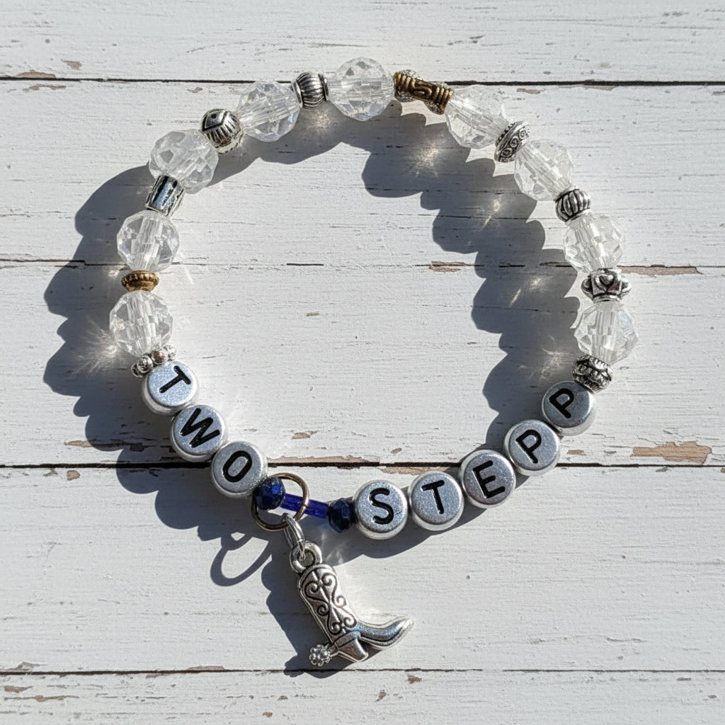 A beaded stretch bracelet with clear faceted beads, silver letter beads spelling 'TWO STEP', small silver spacer beads, a silver cowboy boot charm, and a dark blue cord accent, photographed on a light beige surface with strong shadows.
