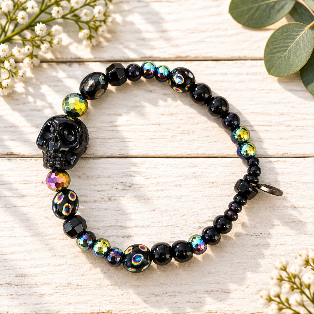 A beaded stretch bracelet featuring a large black skull bead, faceted black beads, and iridescent beads that reflect pink, green, and gold colors, displayed on a light wood grain surface.