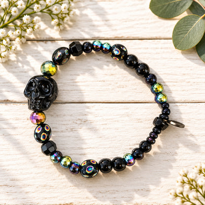 A beaded stretch bracelet featuring a large black skull bead, faceted black beads, and iridescent beads that reflect pink, green, and gold colors, displayed on a light wood grain surface.
