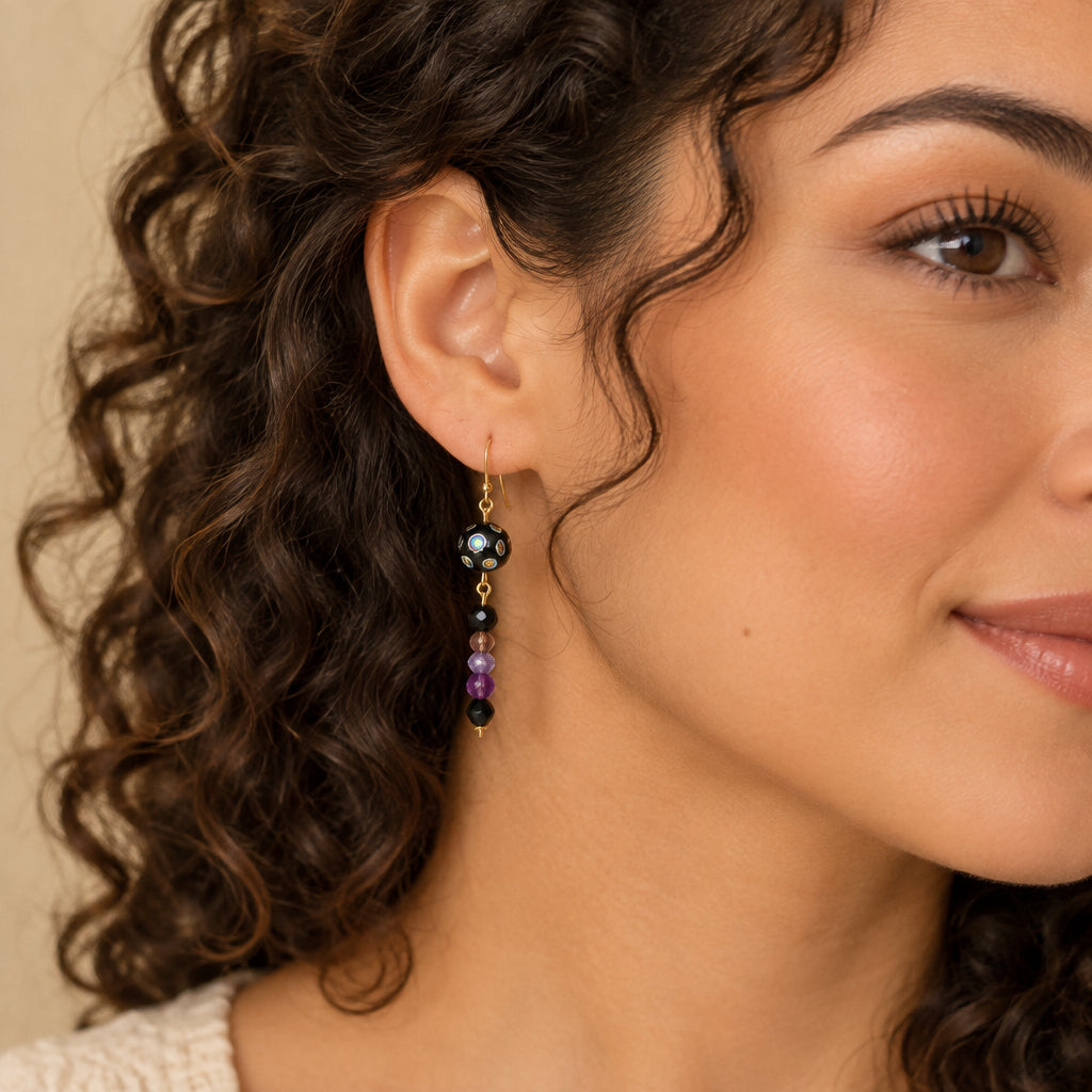 A pair of handmade dangle earrings with gold fishhook wires, black glass beads featuring iridescent colorful spots, and dangling faceted beads in black, brown, and purple, displayed on a woman.