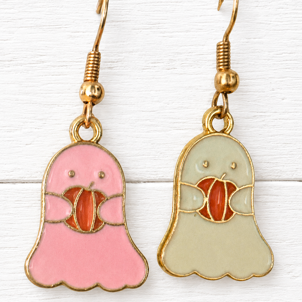 A pair of dangling ghost-shaped earrings with gold-tone hooks. One ghost is pink and holds a red-orange pumpkin, while the other is pale green and holds a red-orange pumpkin.