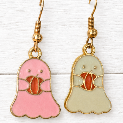 A pair of dangling ghost-shaped earrings with gold-tone hooks. One ghost is pink and holds a red-orange pumpkin, while the other is pale green and holds a red-orange pumpkin.