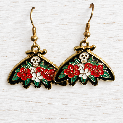 A pair of dangling butterfly-shaped earrings with gold-tone hooks and bodies, decorated with red and white flowers, green leaves, and a small white skull motif on a black enamel background.