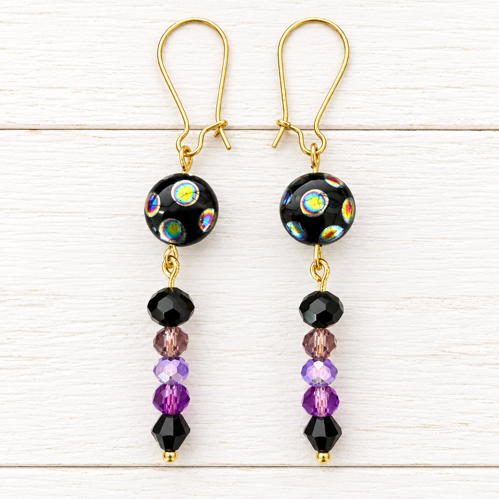 A pair of handmade dangle earrings with gold fishhook wires, black glass beads featuring iridescent rainbow patterns, and dangling purple and black faceted beads, displayed on a white surface.