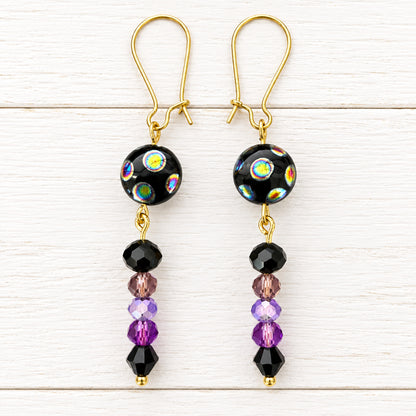 A pair of handmade dangle earrings with gold fishhook wires, black glass beads featuring iridescent rainbow patterns, and dangling purple and black faceted beads, displayed on a white surface.