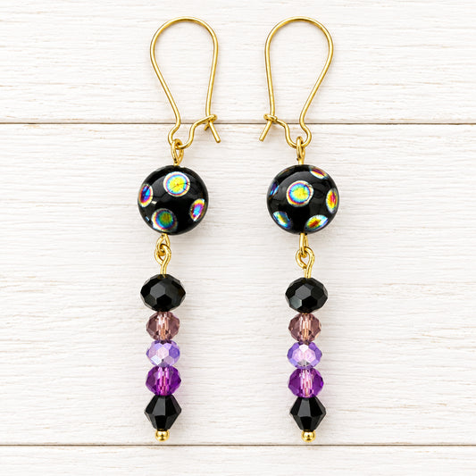 A pair of handmade dangle earrings with gold fishhook wires, black glass beads featuring iridescent rainbow patterns, and dangling purple and black faceted beads, displayed on a white surface.