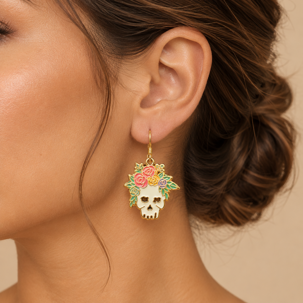 A pair of dangling skull earrings with white enamel skulls, gold-tone metal outlines, and floral crowns in pink, yellow, and green, displayed in a lady's ear.