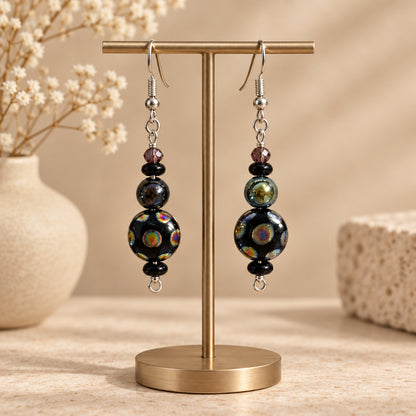 A pair of handmade dangle earrings with black iridescent beads, small dark beads, and silver fishhook wires, displayed on a earring stand 