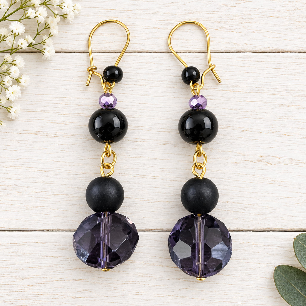 A pair of dangle earrings with gold-tone hooks, small black beads, a tiny purple faceted bead, a larger black bead, and a large faceted dark purple bead at the bottom, displayed on a light wood background.