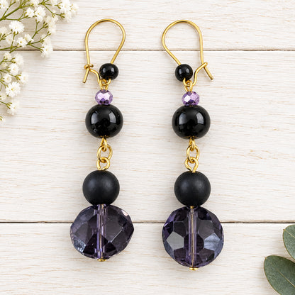 A pair of dangle earrings with gold-tone hooks, small black beads, a tiny purple faceted bead, a larger black bead, and a large faceted dark purple bead at the bottom, displayed on a light wood background.