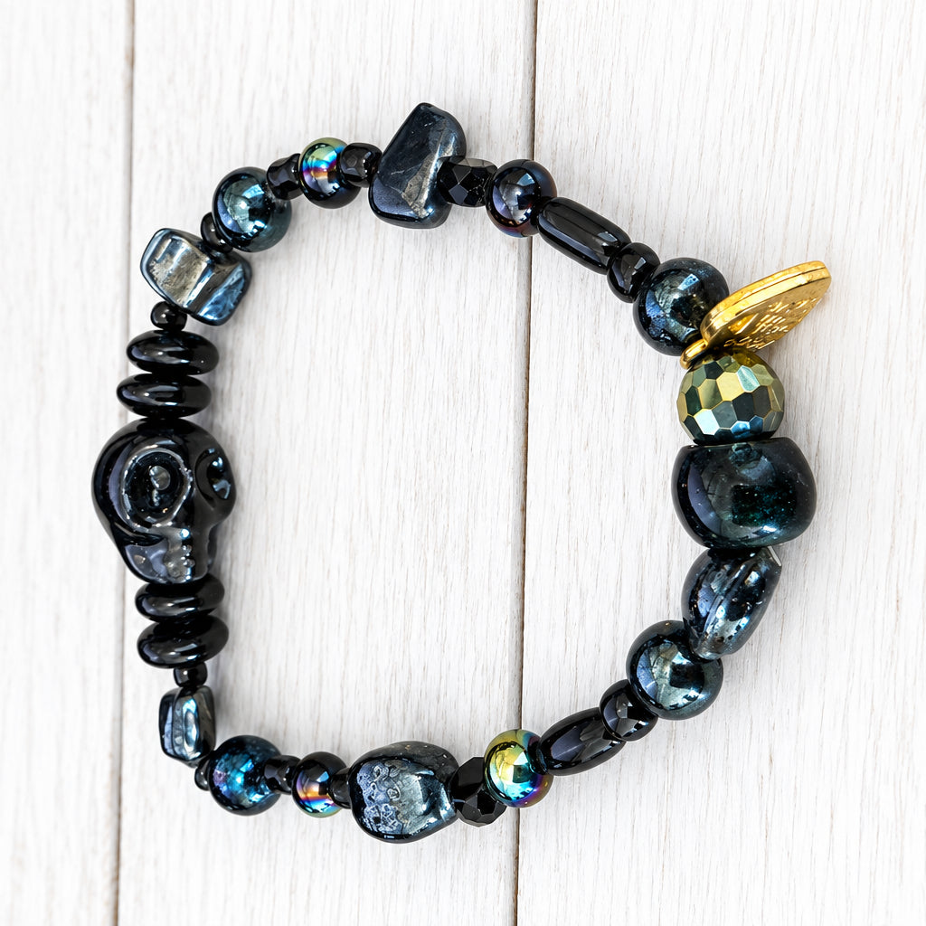 A beaded bracelet with dark polished and faceted beads, a carved black skull centerpiece, and a small gold charm, displayed on a light wood surface.