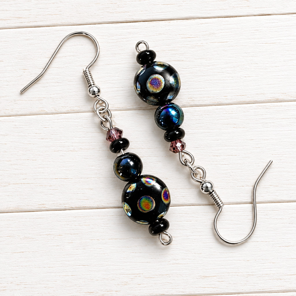 A pair of handmade dangle earrings with dark iridescent beads and silver fishhook wires, displayed on a light wood grain surface.