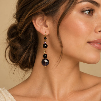 A pair of drop earrings with faceted dark purple beads, matte black beads, small purple accent beads, and gold-tone hooks, displayed on a young lady. 