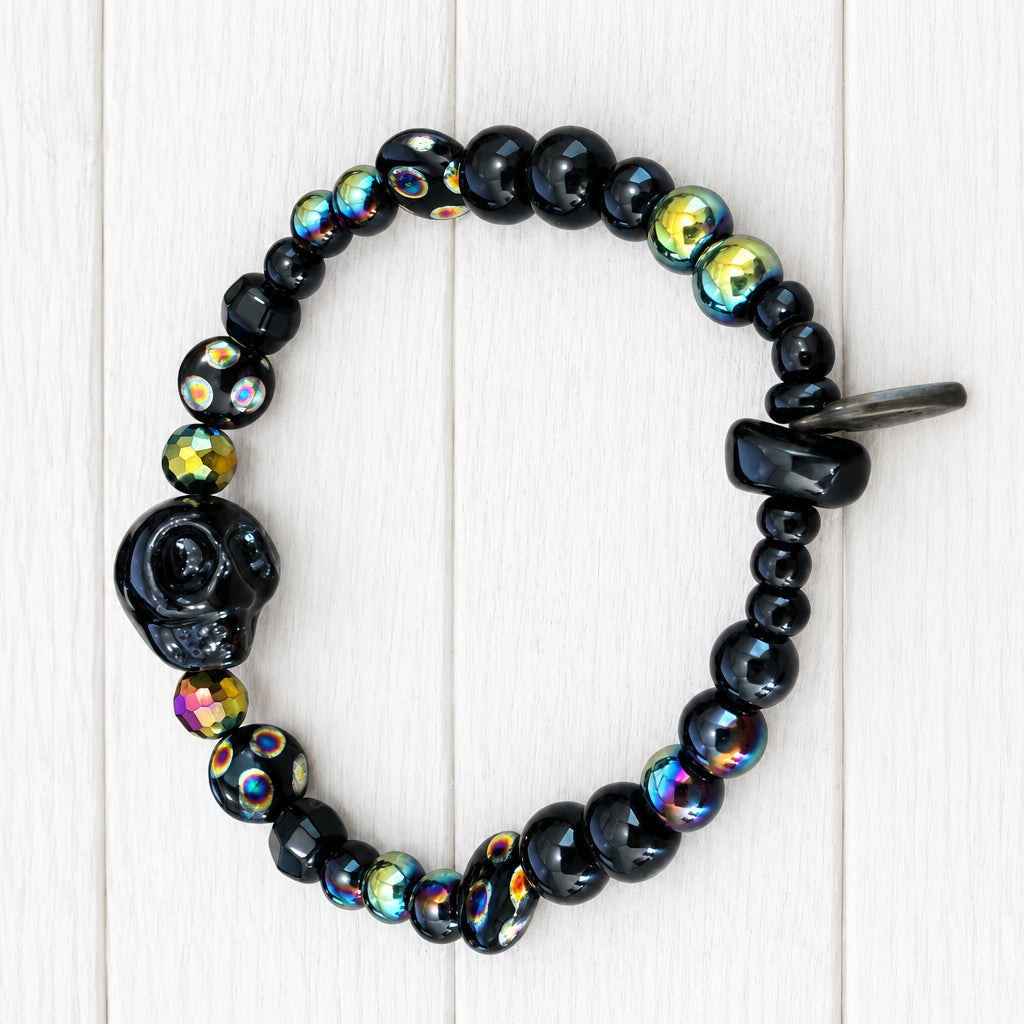 A beaded stretch bracelet featuring glossy black beads, a carved black skull charm, and iridescent beads that reflect gold, green, and purple hues, displayed on a white wood grain surface.