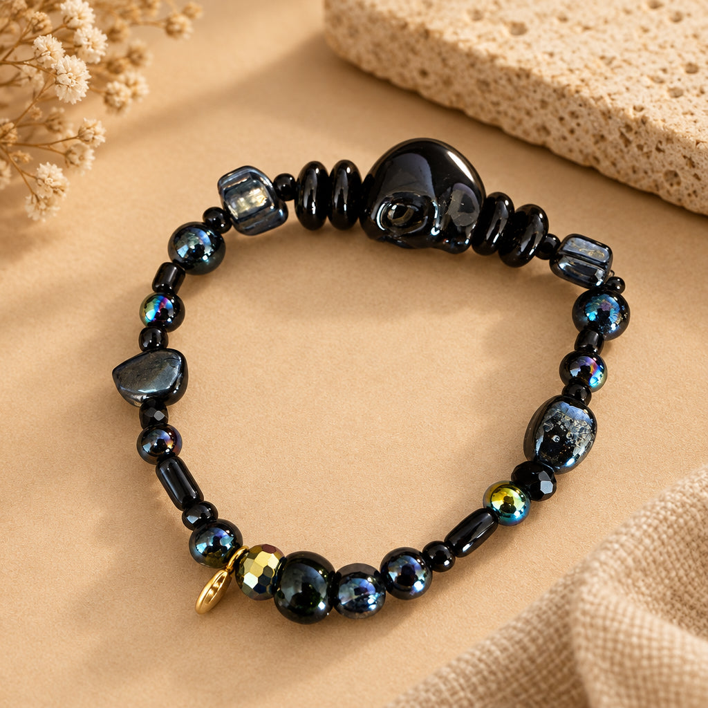 A beaded stretch bracelet made of glossy black beads, polished stone beads, and a few iridescent and gold accent beads, displayed on a light brown surface.