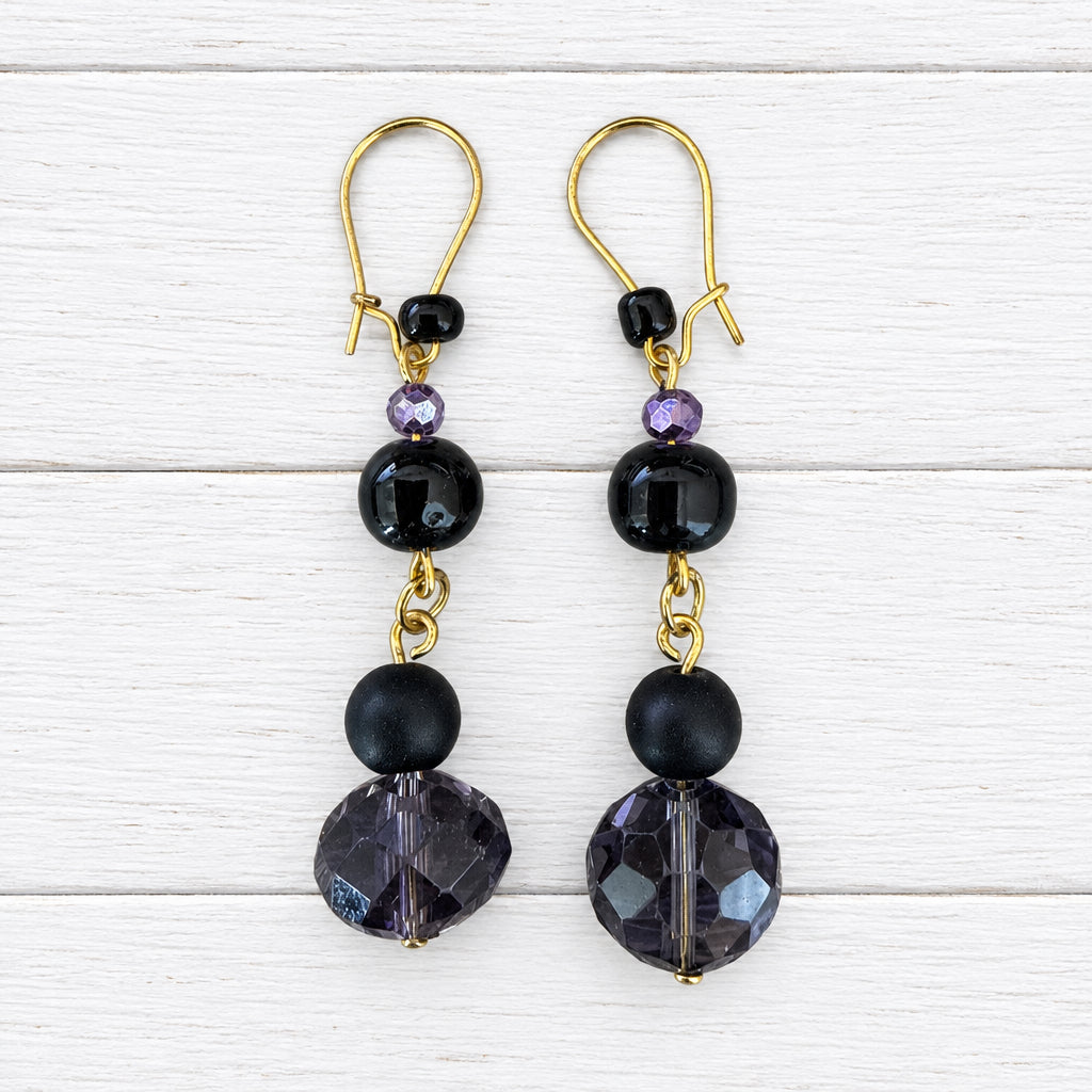 A pair of gold-tone dangle earrings with black faceted beads and dark purple crystal drops, displayed on a light wood grain surface.