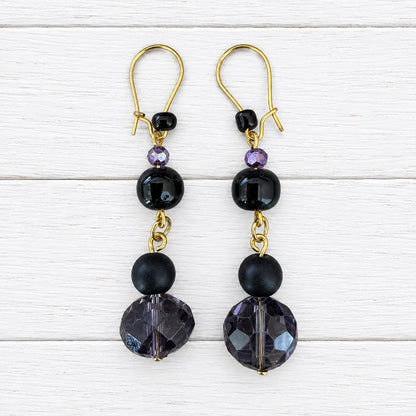A pair of gold-tone dangle earrings with black faceted beads and dark purple crystal drops, displayed on a light wood grain surface.