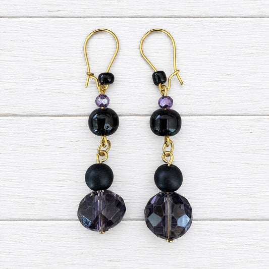 A pair of gold-tone dangle earrings with black faceted beads and dark purple crystal drops, displayed on a light wood grain surface.