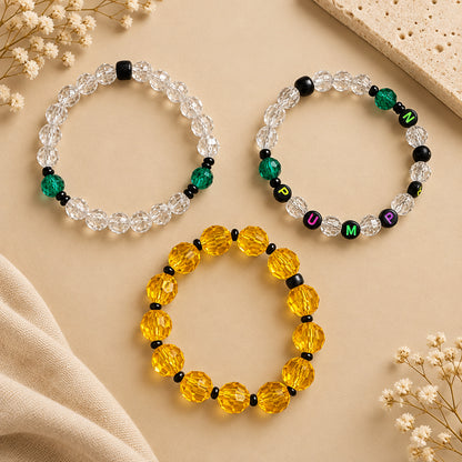 Three beaded stretch bracelets arranged on a light wood tabletop. One bracelet is made of clear faceted beads, one is made of translucent yellow faceted beads, and one is a mix of clear, green, black, and lettered beads spelling out the word pumpkin.