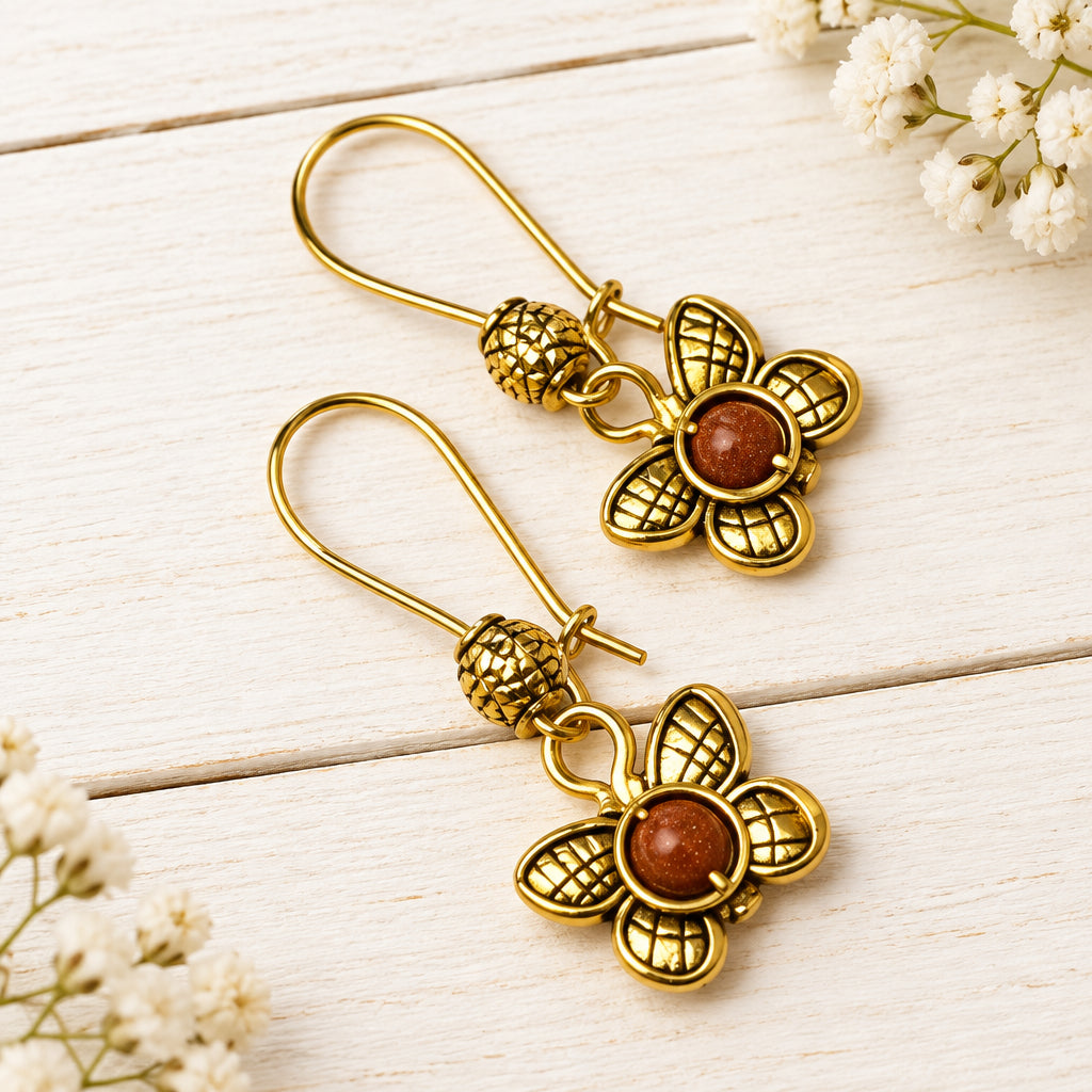 A pair of gold-tone dangle earrings with textured flower charms and round brown stone centers, displayed on a white wooden surface with small white flowers.