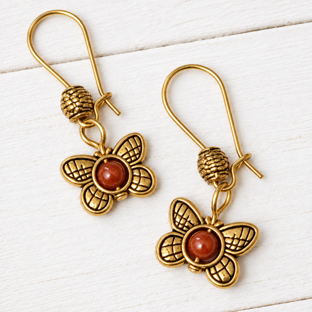A pair of gold-tone butterfly drop earrings with textured bead details and round reddish-brown cabochon centers, displayed on a white wood background.