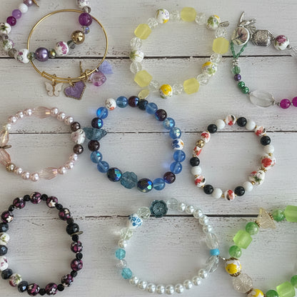 A collection of colorful beaded and pearl bracelets arranged on a light surface with a green floral pattern. The bracelets feature various bead styles, including iridescent blue beads, white pearls, purple beads, yellow beads, and mixed-color floral details.