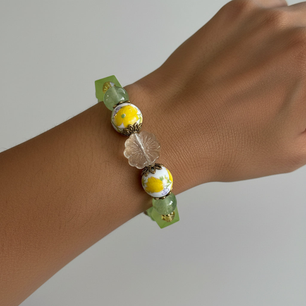A beaded stretch bracelet featuring light green faceted beads, translucent clear beads, and two white glass beads with yellow daisy designs and gold metal spacers, displayed on a neutral light gray background.