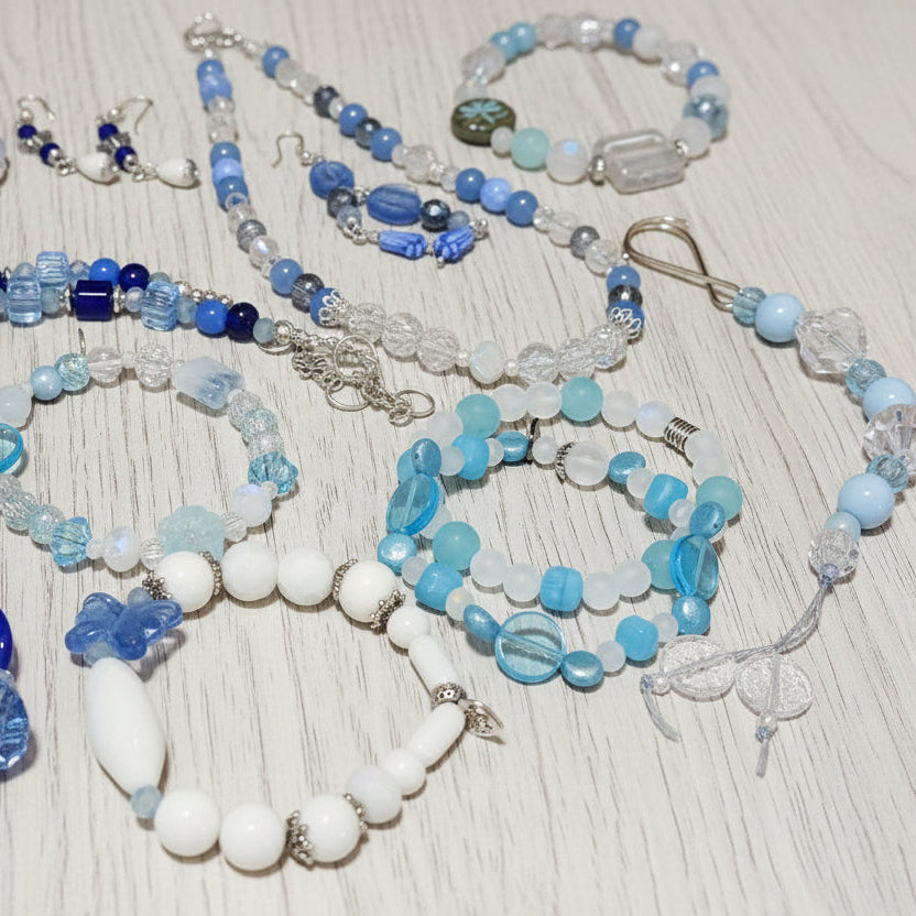 A collection of handmade beaded jewelry, including bracelets, necklaces, and earrings, featuring shades of blue, white, and clear glass beads, arranged on a wooden surface.