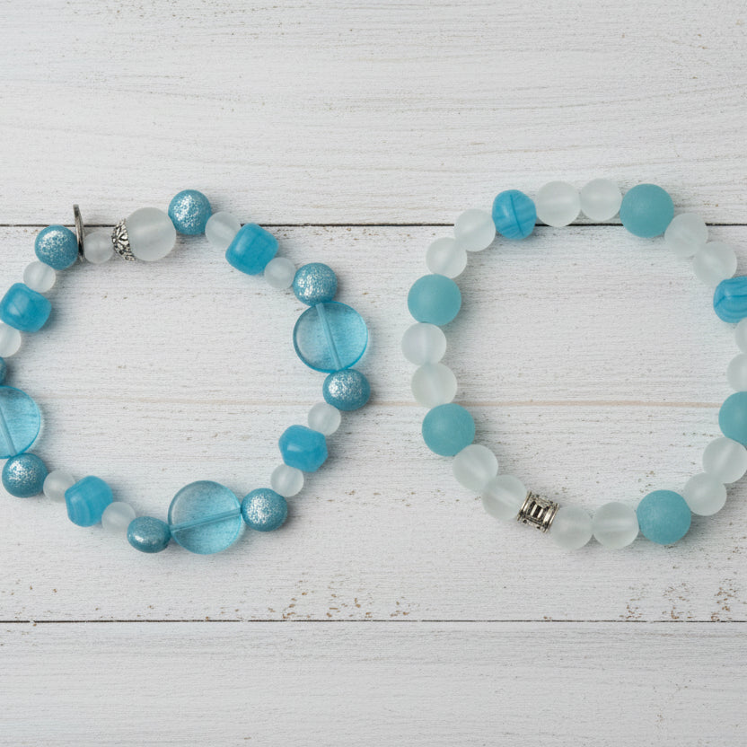 Two beaded stretch bracelets displayed on a light wood plank background. The top bracelet features translucent blue and white round beads with a small silver charm.