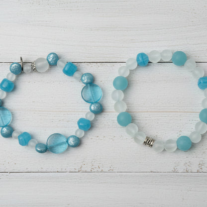 Two beaded stretch bracelets displayed on a light wood plank background. The top bracelet features translucent blue and white round beads with a small silver charm.