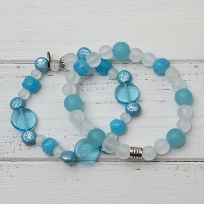 A beaded necklace with round turquoise blue, milky white, and iridescent shell-like beads, accented by silver-toned spacer beads, displayed on a whte wood background.