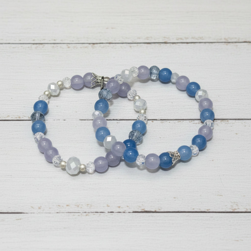 Two beaded bracelets with blue, lavender, and clear faceted beads, displayed on a white-washed wooden surface.