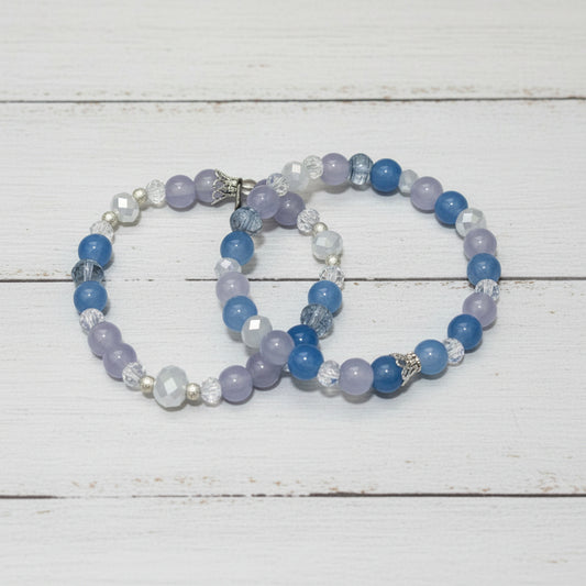Two beaded bracelets with blue, lavender, and clear faceted beads, displayed on a white-washed wooden surface.