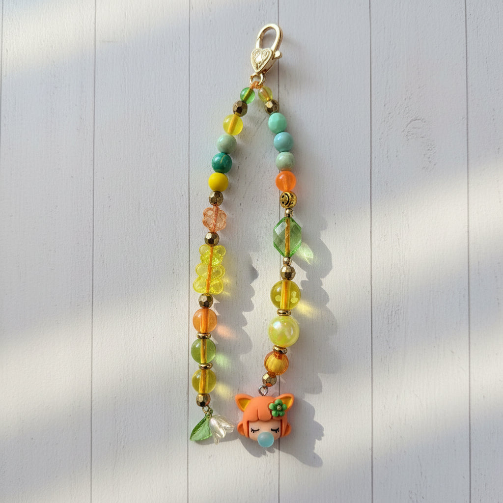 A handmade beaded charm strap with a gold clasp, featuring multicolor beads, a yellow flower, a small green leaf, and an orange fox charm, displayed on a light gray wood plank background.