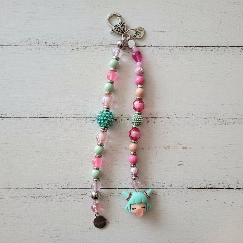 A handmade beaded keychain or phone strap with a silver lobster clasp, featuring pink, mint green, and metallic beads, a green beaded ball accent, a small black charm, and a mint green anime-style character charm at the bottom, displayed against a white wood background.